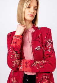 Rdeč cardigan s cvetličnim vzorcem, z gumbom spredaj in rebrastimi manšetami, nošen čez roza srajco s ovratnikom. Mehka tekstura, prilegajoč kroj in vidni vzorci.