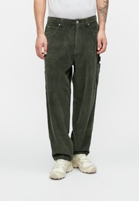 Κορδορέ παντελόνια σε σκούρο πράσινο με χαλαρή εφαρμογή, διαθέτοντας πλαϊνές τσέπες και υφασμάτινο ριγωτό σχέδιο. Φοριούνται με ανοιχτόχρωμα παπούτσια.