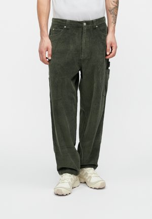 Tummanvihreät kangashousut rentoa leikkausta, joissa on sivutaskut ja teksturoitu ribbikuvio. Yhdistetty vaaleanvärisiin kenkiin.