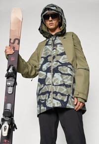 Ski bunda v olivovo zelenej farbe s camo vzorom, s predným zipsom, kapucňou a viacerými vreckami. Model drží lyže s čiernymi viazaniami.