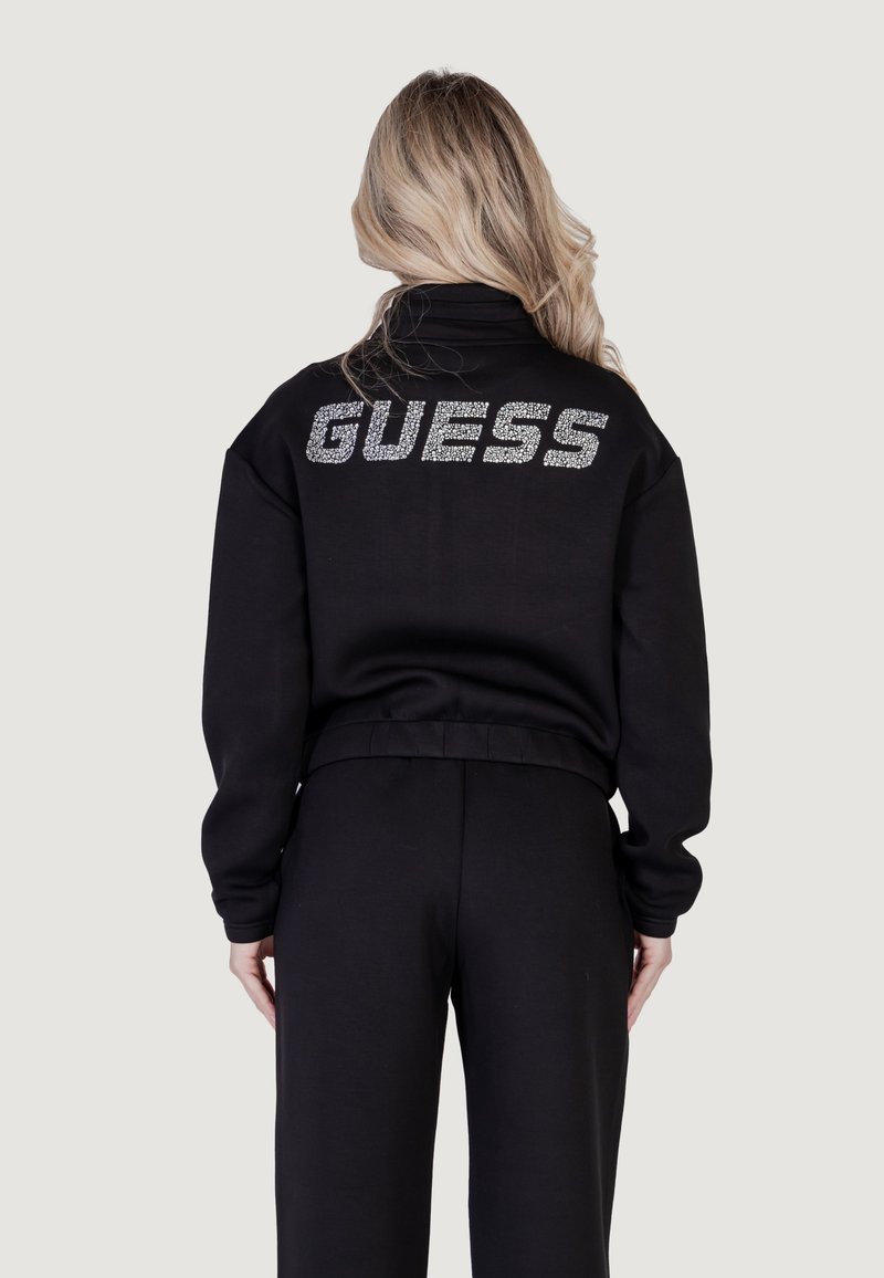 Juodas megztinis su aukšta apykakle ir trumpu dizainu. Nugarėlėje puikuojasi žvilgantis "GUESS" logotipas, padengtas cirkoniais.
