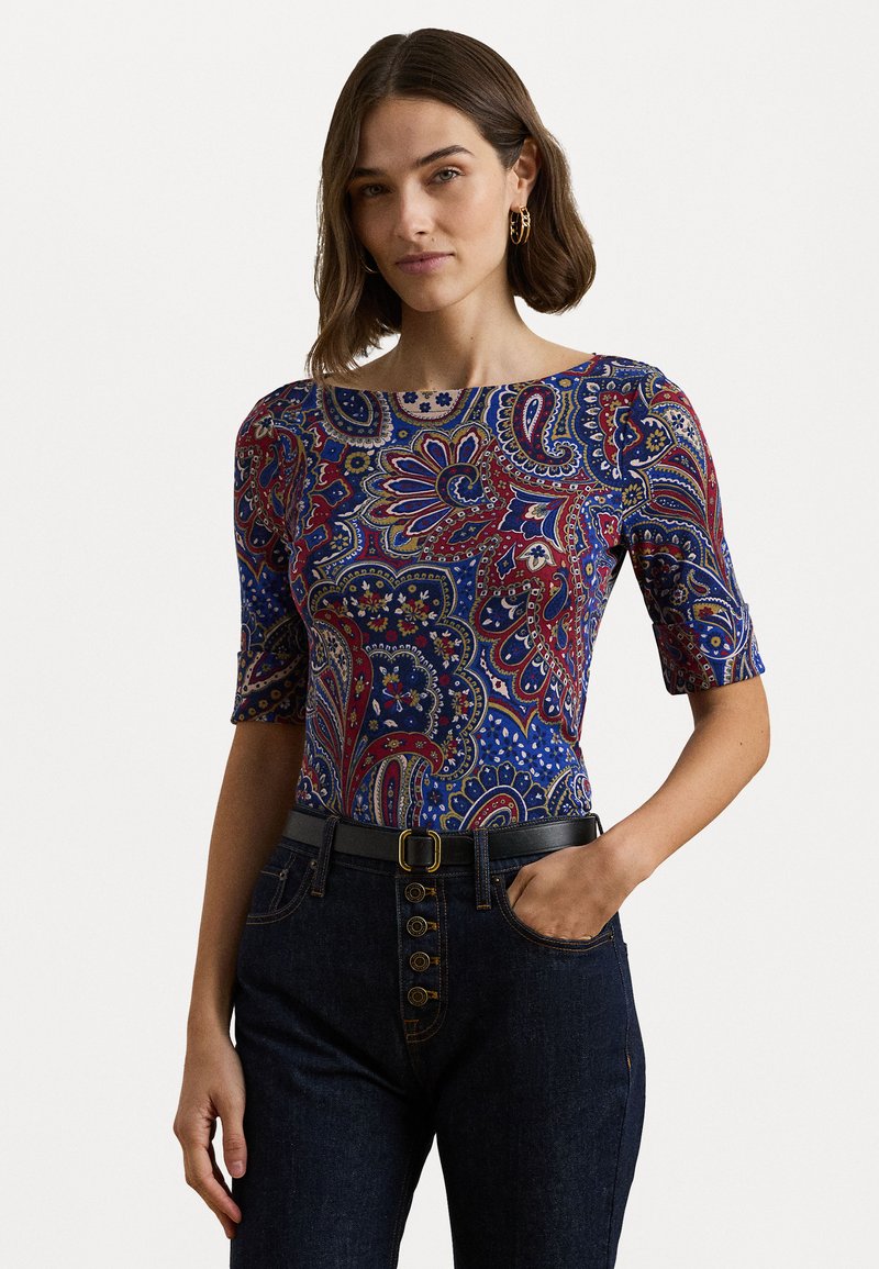 Top con motivo a paisley blu, rosso e crema; silhouette aderente; scollo rotondo; maniche corte; indossato con jeans scuri e una cintura nera.