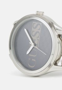 Silberne Analog-Uhr mit grauem Zifferblatt, großem "GUESS"-Logo, markanten Zeigern und strukturiertem Ziffernrand. Zirkulare Gehäuse aus Edelstahl.