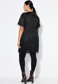 Abito nero con frange, maniche corte, tessuto trasparente e taglio fino al ginocchio. Abbinato a leggings neri e stivaletti alla caviglia bordeaux lucidi.