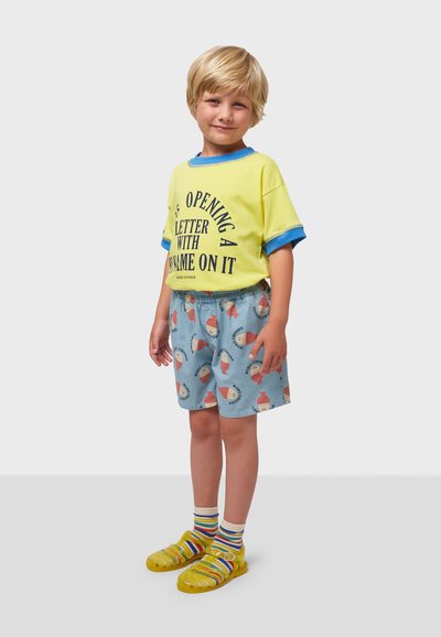 Camiseta amarilla con detalles azules y texto impreso, combinada con pantalones cortos azules que tienen un patrón de conos de helado. Sandalias amarillas y calcetines a rayas.