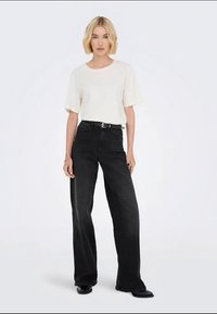 T-shirt branco de manga curta combinado com jeans pretos de perna larga de cintura alta. Os jeans apresentam um cinto e um acabamento liso e texturizado.
