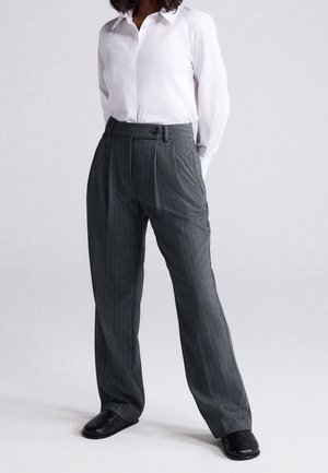 Kvinde iført hvid skjorte med krave, grå høj talje brede bukser med nålestriber og sorte flettede loafers, stående med hænderne bag ryggen.