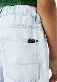 Pantalones de mezclilla azul claro con cinturilla fruncida, bolsillo trasero y una etiqueta negra "Garcia" cosida en el lado.