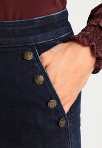 Gonna di jeans scura con vita aderente, caratterizzata da cinque bottoni decorativi in ottone sul lato, e una mano infilata nella tasca.