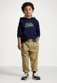 Jaunas berniukas su garbanotais plaukais, apsirengęs tamsiai mėlynu polo stiliaus džemperiu, smėlio spalvos kelnėmis, kurių apačia apversta, ir tamsiai mėlynomis sportinėmis bateliais su baltais raišteliais, stovi prieš paprastą foną.