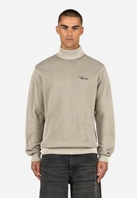 Jasno beżowy sweter z wysokim kołnierzem, prążkowany, z długimi rękawami i dopasowanym dołem, z małym czarnym logo na piersi. Tekurowana tkanina.