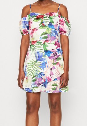 Vrouw draagt een witte, bloemige off-shoulder jurk met gerimpelde zoom en groene, roze, blauwe en paarse tropische bloemenpatronen.