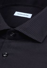 Chemise habillée noire texturée avec col plié, deux boutons noirs et une étiquette blanche « seidensticker » à l'intérieur du col.