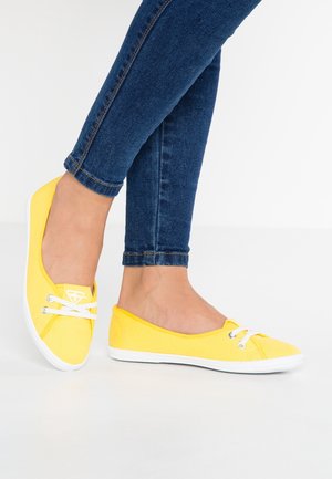 Żółte płócienne buty wsuwane z białymi sznurówkami i gumowymi podeszwami, w połączeniu z niebieskimi wąskimi dżinsami, mają okrągły czubek i minimalne logo.