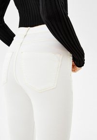 Jeans blancos de tejido suave y elástico, con dos bolsillos traseros y costuras en contraste color crema. Combinados con una blusa negra de canalé.