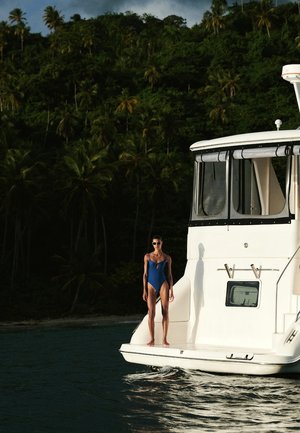 Sininenuinen nainen sinisessä uimapuvussa seisoo valkoisen veneen taka-alustalla trooppisen metsän rannalla, jossa kasvaa palmupuita.