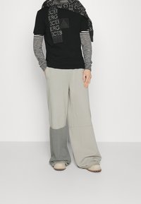 Camiseta negra de manga corta con logo, camiseta interior de manga larga a rayas grises, pantalones holgados grises y gris claro, y zapatos beiges sin cordones.