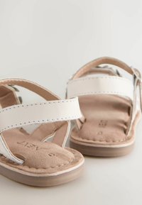 Sandali bianchi in pelle con cinturini incrociati, plantare in suede marrone, suola color beige chiaro e dettagli di cucitura sui cinturini.
