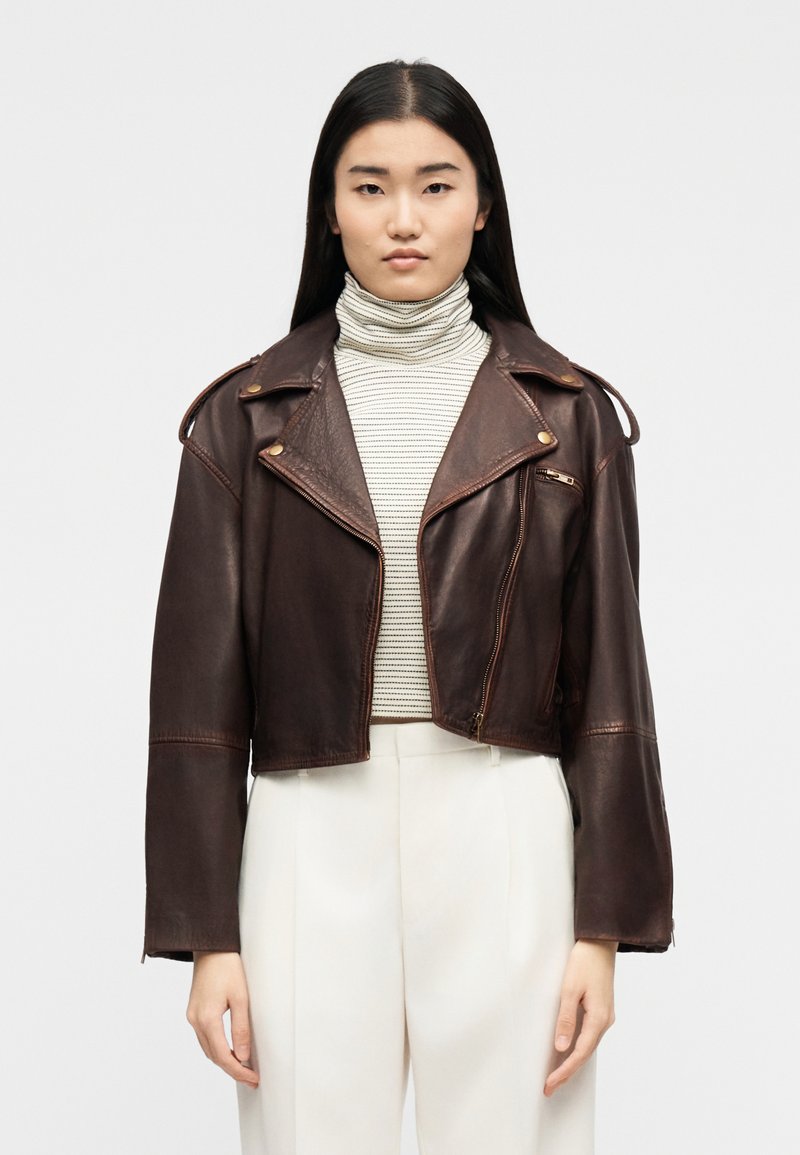 Γυναίκα με σκούρο καφέ κοντό δερμάτινο μπουφάν πάνω από μπεζ ριγέ ζιβάγκο και λευκό παντελόνι ψηλής μέσης, στεκόμενη μπροστά σε μονόχρωμο φόντο.