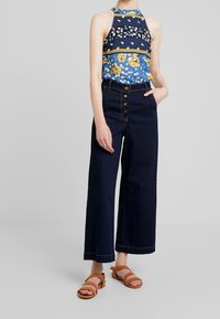 Kvetinový top na holá ramena v modrých a žlutých tónech; tmavé džíny s vysokým pasem; hnědé sandály; zapínání na knoflík; úzký korzet a široké nohavice.