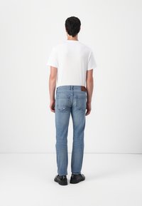 Helle blaue Jeans mit geradem Bein, mit einem braunen Lederpatch am hinteren Bund und zwei Gesäßtaschen. Getragen mit einem weißen T-Shirt.