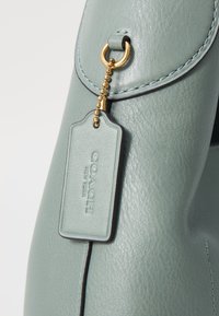 Borsa a mano in pelle verde acqua con una superficie liscia, caratterizzata da dettagli in metallo dorato e un'etichetta impressa con "COACH NEW YORK."