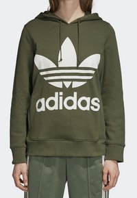 Olivgrön hoodie med en stor vit Adidas-logotyp och trebladig design. Har knytband i huvan och ribbade muddkanter.