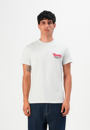 Uomo con capelli neri corti che indossa una maglietta grigia chiaro di Tommy Jeans e jeans blu scuro, in piedi contro uno sfondo bianco.