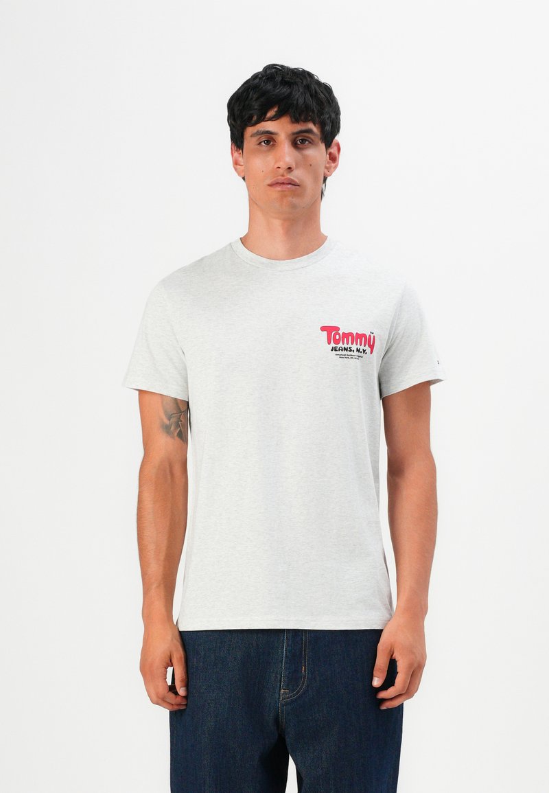 Homme avec les cheveux noirs courts portant un t-shirt gris clair de Tommy Jeans et un jean bleu foncé, se tenant contre un fond blanc.