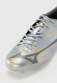 Silver soccer cleat with grey laces, featuring a textured finish, gold stripes, and a prominent logo on the side.