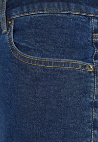 Jean en denim bleu, avec une poche arrière standard, des coutures dorées et une texture lisse. Le tissu présente un léger effet délavé.