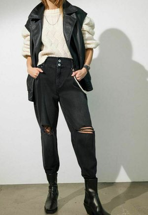 Schwarze zerrissene Jeans kombiniert mit einem strukturierten cremefarbenen Strickpullover und einer ärmellosen schwarzen Lederjacke; klobige schwarze Stiefeletten runden den Look ab.