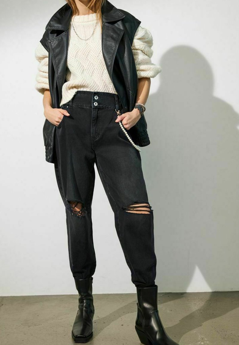 Svarta ripped jeans i kombination med en texturerad krämfärgad stickad tröja och en svart läderväst; chunky svarta ankelkängor fulländar looken.