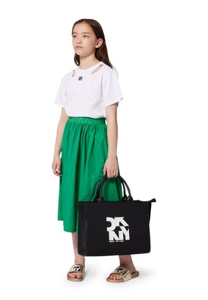 Sac fourre-tout noir avec le logo blanc DKNY, finition brillante, avec deux poignées supérieures. Le modèle porte un T-shirt blanc, une jupe verte et des sandales dorées à glisser.