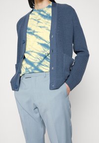 Blå stickad cardigan med texturerade mönster, som har två framfickor och knapplåsingar, över en gul tie-dye skjorta och ljusa byxor.