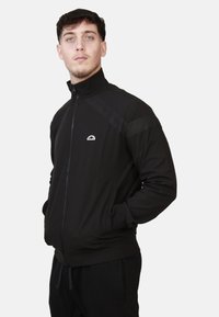 Giacca nera con zip e colletto alto, tasche laterali e sottili strisce sulle spalle. Presenta un piccolo logo sul petto. Tessuto dalla superficie liscia.