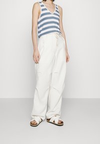 Persona che indossa un top senza maniche a righe bianche e blu, pantaloni bianchi larghi con vita regolabile e sandali bianchi con cinturini incrociati.