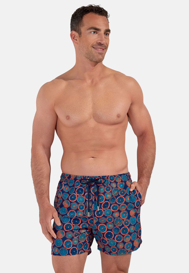 Mann mit kurzen lockigen Haaren, der blaue Badeshorts mit orangefarbenen und roten Zitrusfruchtmustern trägt und mit einer Hand in der Tasche steht.