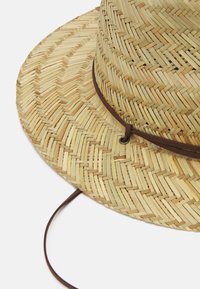 Oljenna aurinkohattu leveällä lierillä, luonnollinen beige väri, kudottu rakenne ja ruskea nahkaremmii, joka on pujotettu metallirenkaaseen.