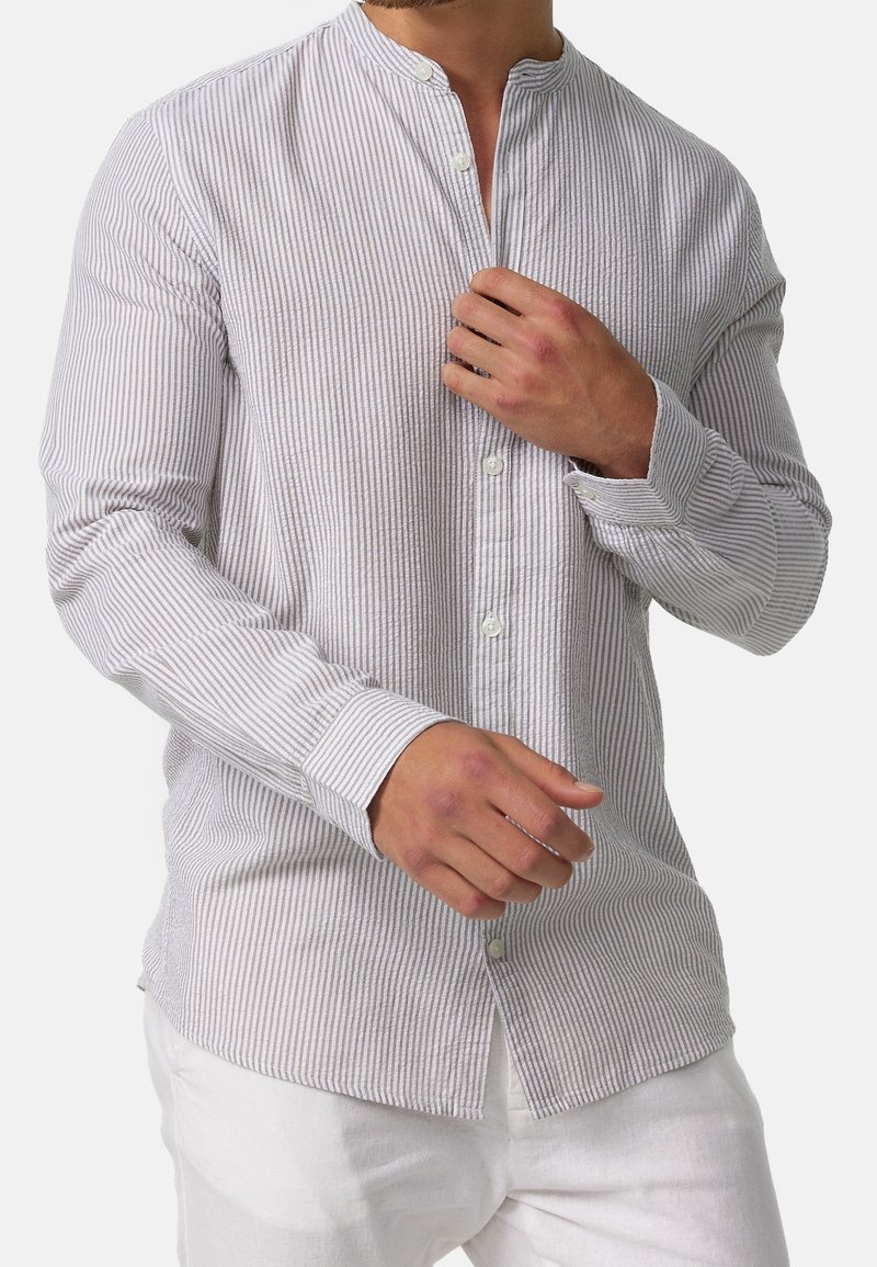 Homme portant une chemise à rayures verticales grises claires et blanches avec col mao, manches longues et pantalon blanc, ajustant le devant de la chemise.