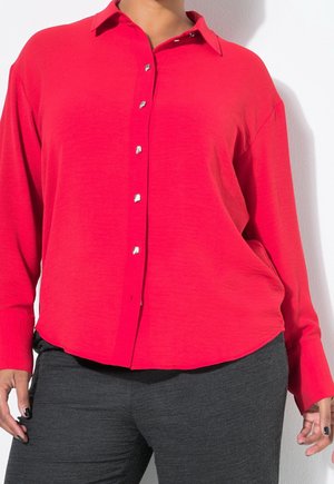 Personne portant une chemise rouge vif à boutons argentés et un pantalon gris foncé, debout devant un fond blanc uni.