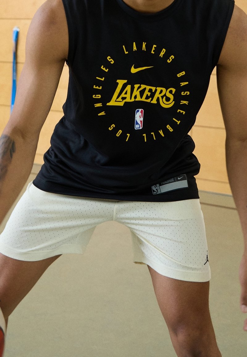 Personne portant un maillot sans manches noir des Los Angeles Lakers et un short de basket blanc, prête en position défensive à l'intérieur.