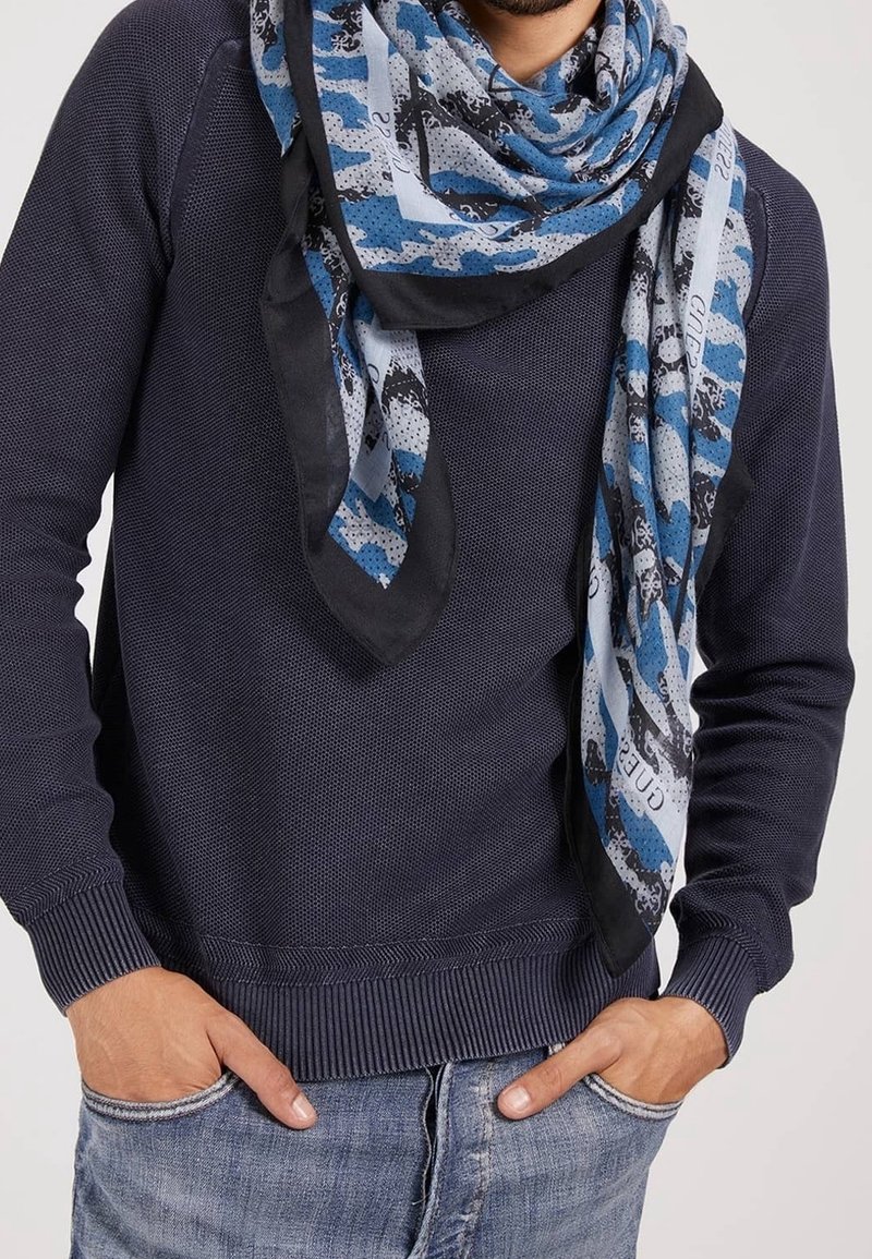 Homme portant un pull texturé bleu foncé et une écharpe bleue à motifs, les mains dans les poches d'un jean bleu clair, sur fond uni.