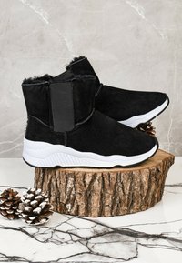 Botas de tobillo negras con material tipo ante, suela de goma blanca y paneles laterales elásticos. Parte superior texturizada en negro con borde de suave pelo.