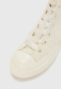 Høyhælte sneakers i kremfarge med snøring, dekke av blonder, satengsnører og en glanset tåhette med strukturert gummisåle.