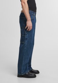 Zilas denim bikses ar taisno kāju piegriezumu, kurām ir vairākas kabatas un izturīga tekstūra. Valkātas ar melniem slip-on apaviem.