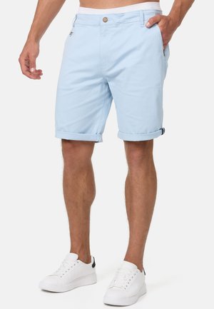 Mand iført lyseblå shorts med opkørte kanter og hvide sneakers, stående med den ene hånd i lommen mod en ensfarvet baggrund.
