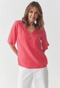 Korallivärvi blouse avatud kaelusega ja lilleline tikand kaeluse ümber. Pehme kangas, lühikesed varrukad ja vabalt istuv lõige, koos valgete pükstega.