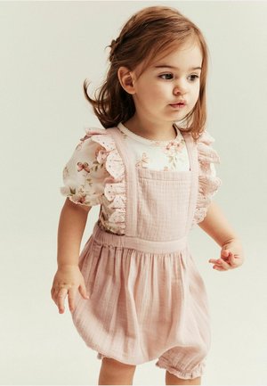 Kleinkind-Mädchen mit hellbraunen Haaren, das ein geblümtes Oberteil mit Schmetterlingsmuster und ein rosa Kleid mit Rüschen trägt, steht und schaut nach rechts.