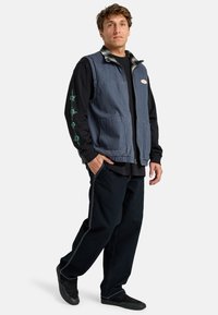 Denim vest in dark blue with a zipper, two front pockets, worn over a black long-sleeve shirt. Black pants with white stitching, completes the outfit.